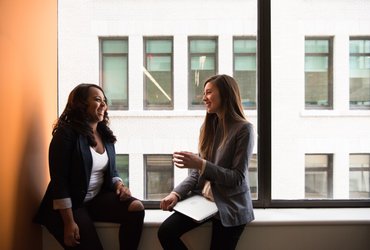 IWCE Be part of the community - Two women sitting on windowsill chatting  - Photo by Christina Wocintechchat on Unsplash